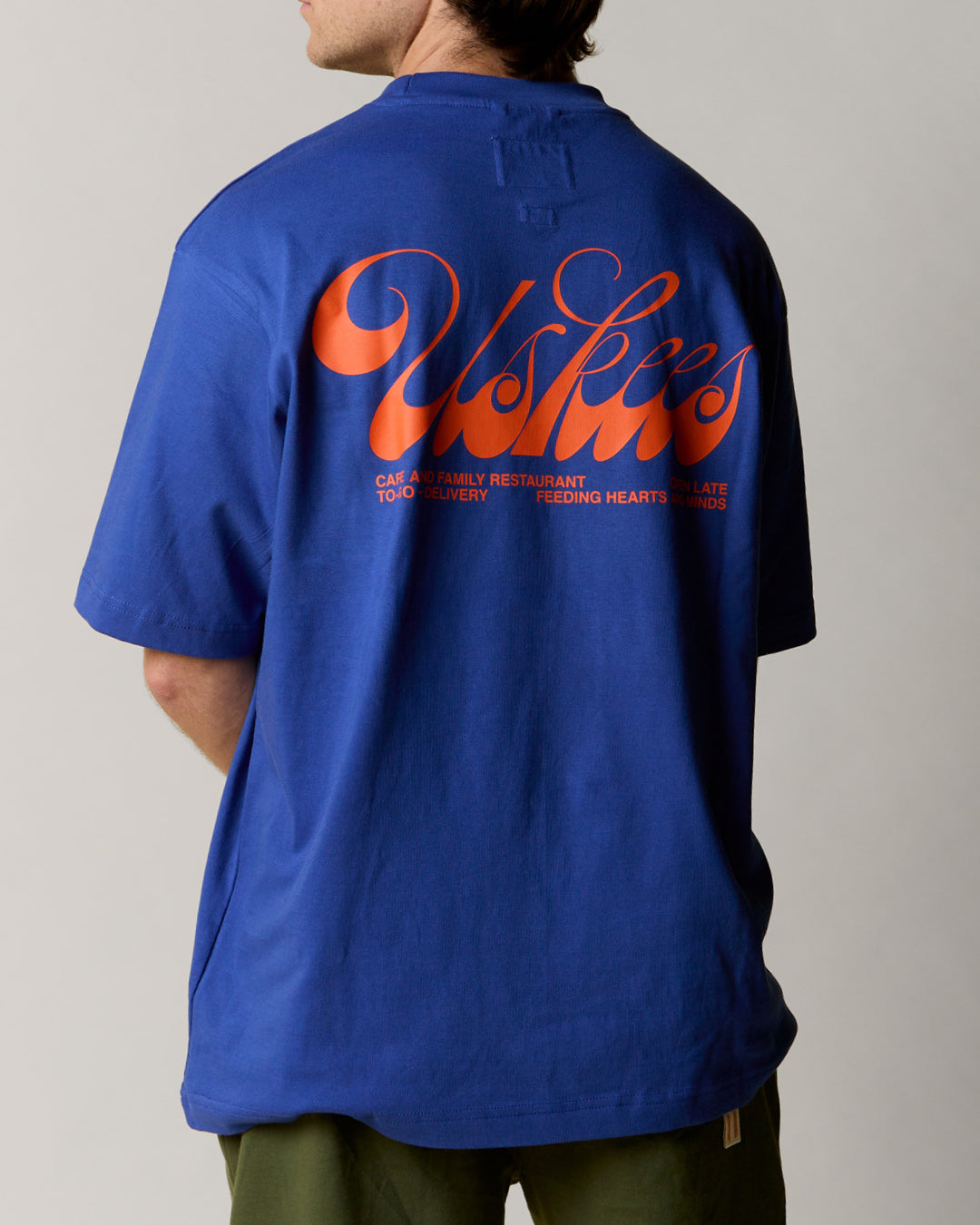 Reverse view of man wearing the uskees ultra blue men's 'diner' tee, demonstrating the oversized fit and the large, bright orange 'diner' logo displayed across the back.