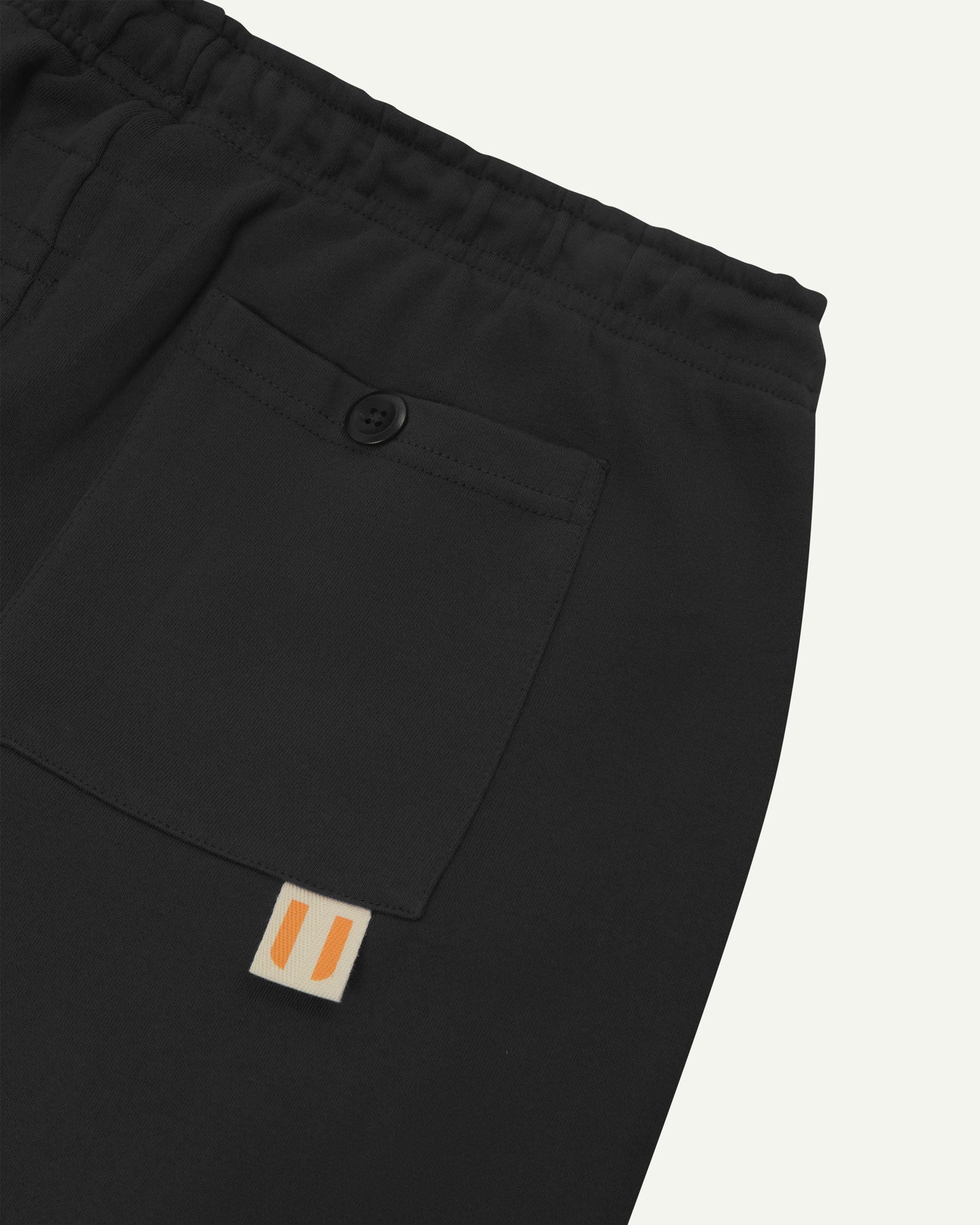 Back close view of faded black organic cotton joggers by Uskees showing the buttoned back pocket and logo label.