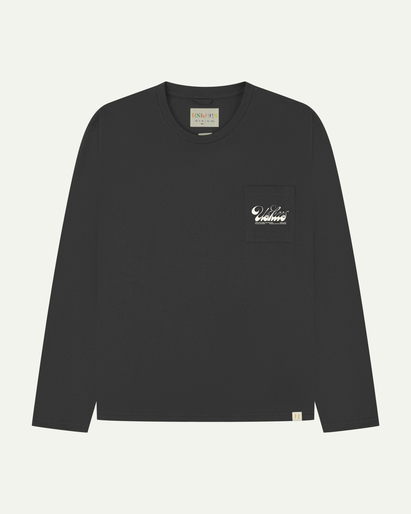 Full flat view of faded black organic cotton Uskees long-sleeve T-shirt for men, showing the small white 'diner' logo on the breast pocket and the Uskees branding labels on the neck and hem.