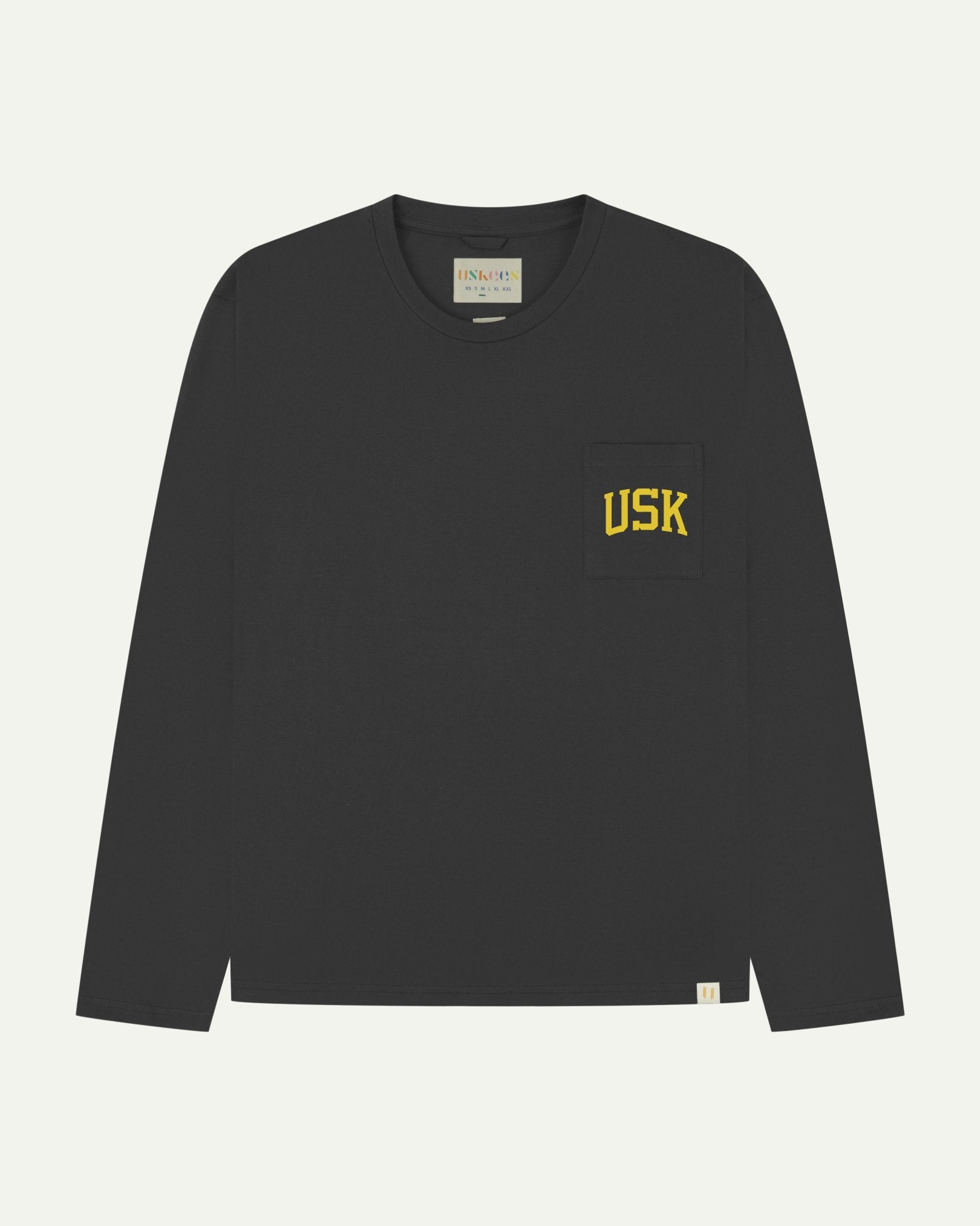 Full flat view of faded black organic cotton signature long sleeve T-shirt for men, showing the varsity inspired 'USK' logo in yellow on the chest pocket, and the Uskees branding labels on the neck and hem.