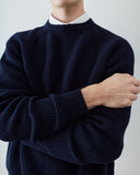 A close-up (neck to waist) of a man wearing a navy blue ribbed knit sweater over a white collared shirt, with his arms crossed.