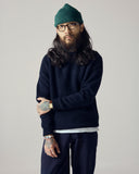 Male model wearing an Uskees midnight blue ribbed sweater, green wool beanie, and blue trousers against a plain background.