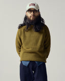 Male model wearing an Uskees woodland green sweater, a cream signature cap and midnight blue trousers.