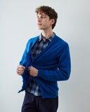 A man wearing a vibrant blue knit cardigan over a blue and white plaid shirt, paired with dark trousers, standing against a plain light gray background.