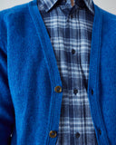 A close-up (neck to waist) of a model wearing a bright blue knit cardigan worn over a blue and white plaid button-up shirt. The cardigan has dark brown buttons and is shown against a plain light gray background.