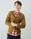 A man wearing a textured green-brown cardigan over a red and white plaid shirt, paired with navy blue pants, photographed against a light gray studio background.