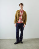 A man stands in a minimalist studio setting wearing an Uskees green/brown knit cardigan over a red and white plaid shirt, paired with navy blue trousers and black leather shoes.