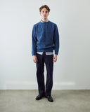A man wearing an Uskees fluffy textured blue knit sweater over a striped collared shirt, paired with dark navy trousers and black leather shoes, standing against a plain light gray wall.