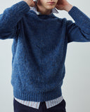 A man wearing a fluffy textured blue knit sweater over a striped collared shirt and dark trousers, shown from the chest down to the waist against a plain light gray background.