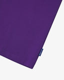 View of hemline of Uskees purple cotton T-shirt with focus on cotton jersey fabric and blue 'USKEES' logo tab on the seam.