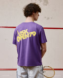 Back view of model wearing the Uskees purple organic cotton T-shirt for men, showing the loose fit and bold 'USK Open 79' brand graphics.