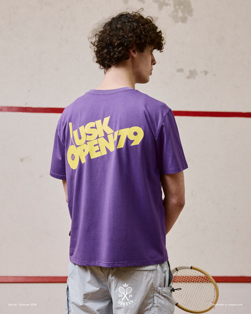 Back view of model wearing the Uskees purple organic cotton T-shirt for men, showing the loose fit and bold 'USK Open 79' brand graphics.