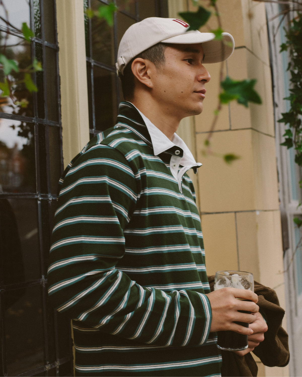 A man wearing an Uskees green and white striped long-sleeve rugby shirt, a cream baseball cap, and holding a drink, standing outdoors next to a building.