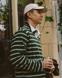 A man wearing an Uskees green and white striped long-sleeve rugby shirt, a cream baseball cap, and holding a drink, standing outdoors next to a building.