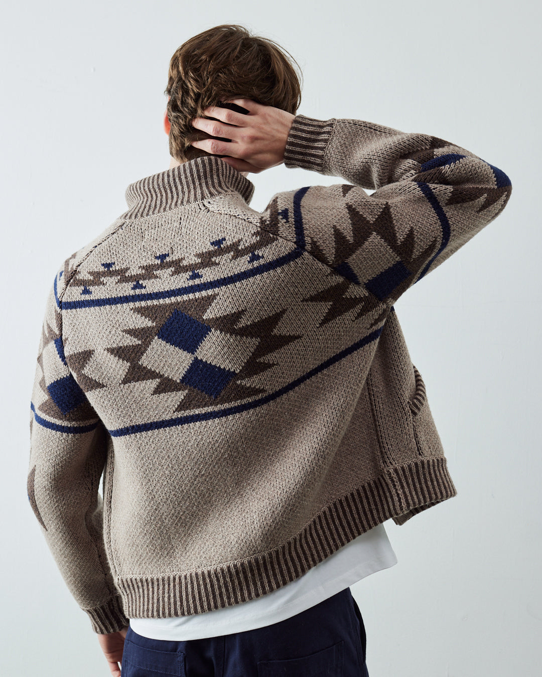 A male model viewed from behind wearing an Uskees beige knit cardigan with a dark brown and navy blue geometric pattern on the back, paired with a white t-shirt and navy pants.