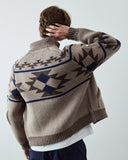 A male model viewed from behind wearing an Uskees beige knit cardigan with a dark brown and navy blue geometric pattern on the back, paired with a white t-shirt and navy pants.