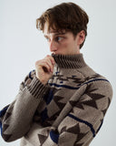 Front view of a man wearing an Uskees  beige knit turtleneck sweater with a dark blue and brown geometric pattern, posing against a light gray background.