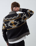 A male model viewed from behind wearing a black knit cardigan with a geometric pattern in white and mustard yellow, layered over a white t-shirt and black trousers, against a light gray background.