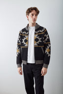 A male model wearing an Uskees black knit zip-up cardigan with a geometric pattern in grey and mustard yellow, layered over a white t-shirt and black trousers, standing against a light gray studio background.