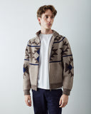 A male model wearing an Uskees beige knit zip-up cardigan with a brown and navy blue geometric pattern, layered over a white t-shirt and dark navy pants, set against a plain light gray studio background.