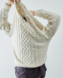 A person wearing a chunky cream cable-knit sweater pulling it off over his head. The sweater features intricate Celtic-style cable patterns and ribbed hem, paired with dark plaid shirt against a light gray background.
