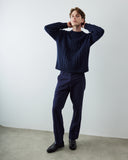 A male model wearing an Uskees navy blue cable-knit sweater and matching navy trousers, standing against a plain light gray wall with a concrete floor.