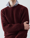 A close-up (neck to waist) of a person wearing an Uskees chunky knit crew neck sweater in deep burgundy, layered over a striped collared shirt, against a plain light gray background.