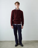A man stands against a plain light gray wall wearing an Uskees burgundy ribbed knit sweater over a light blue and white striped collared shirt, navy trousers, and black leather shoes.