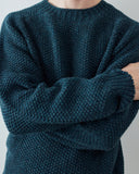 A close-up (neck to waist) of a person wearing a dark teal textured knit sweater with crossed arms, showing the ribbed collar and cuffs against a plain light gray background.