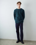 A man wearing an Uskees dark teal knit crew neck sweater, navy blue trousers, and black leather loafers, standing against a plain white wall.