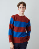 A man wearing a crewneck knit sweater with horizontal stripes in rust red and royal blue, paired with dark navy trousers, standing against a light gray studio background.