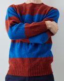 Close-up (neck to waist) of a person wearing a chunky knit sweater with wide horizontal stripes in rust red and bright blue, standing with arms crossed against a plain light gray wall.