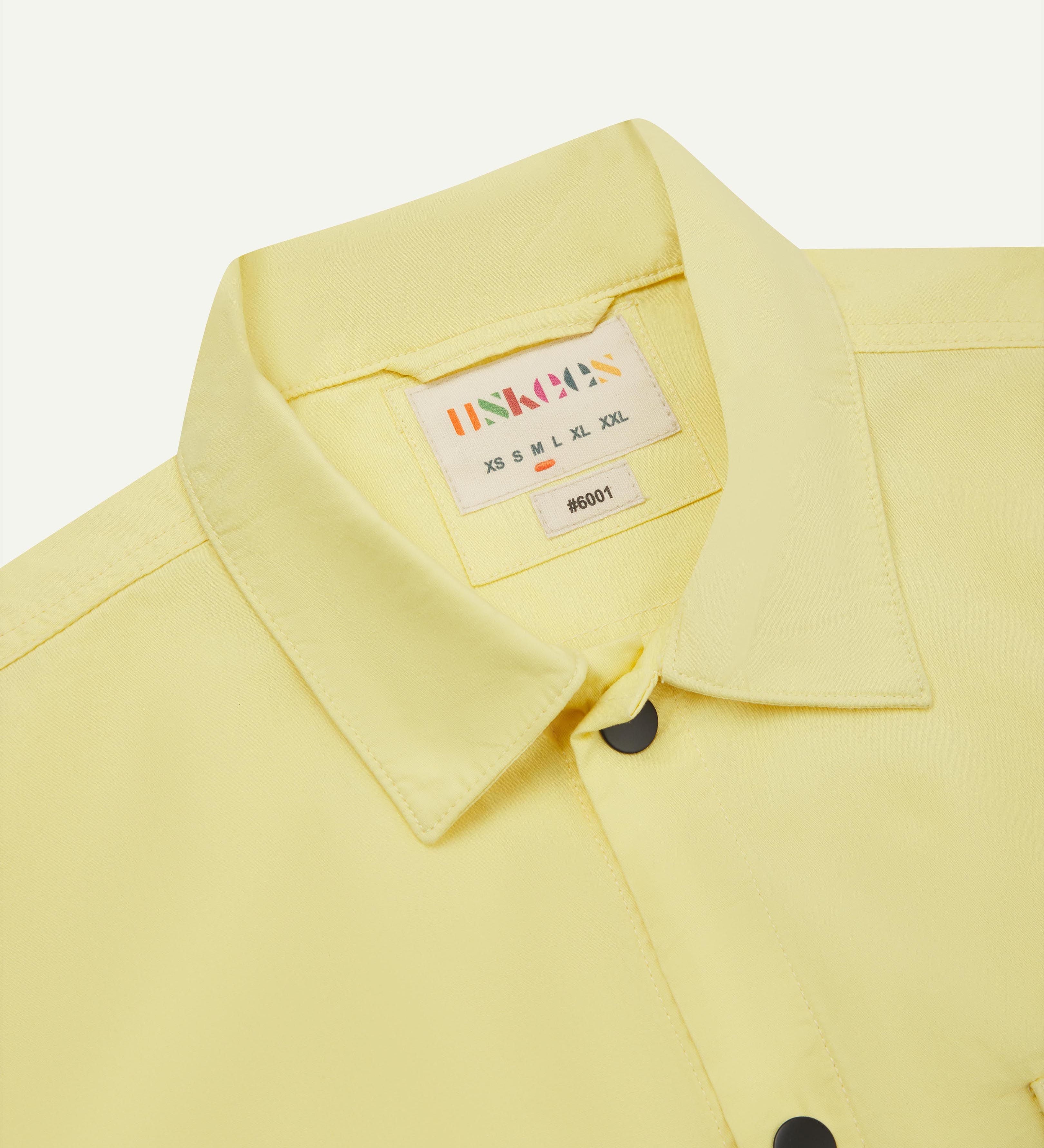 Front close-view of pale yellow buttoned lightweight overshirt from Uskees, showing black press studs and neck label