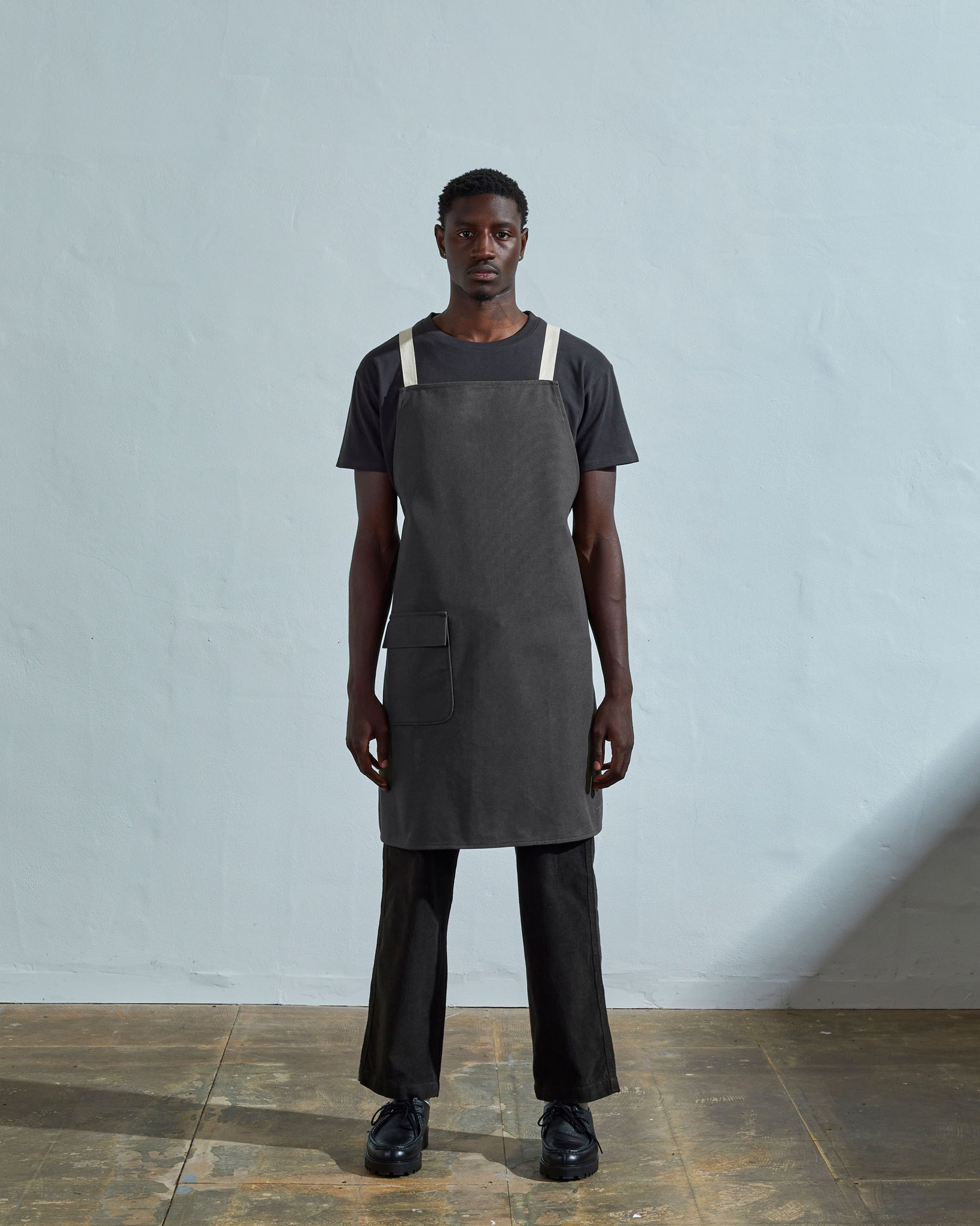 Full-length, front view of male model wearing Uskees #9001 charcoal-grey canvas work apron by Uskees. Showing convenient hip pocket and contrasting cream straps.