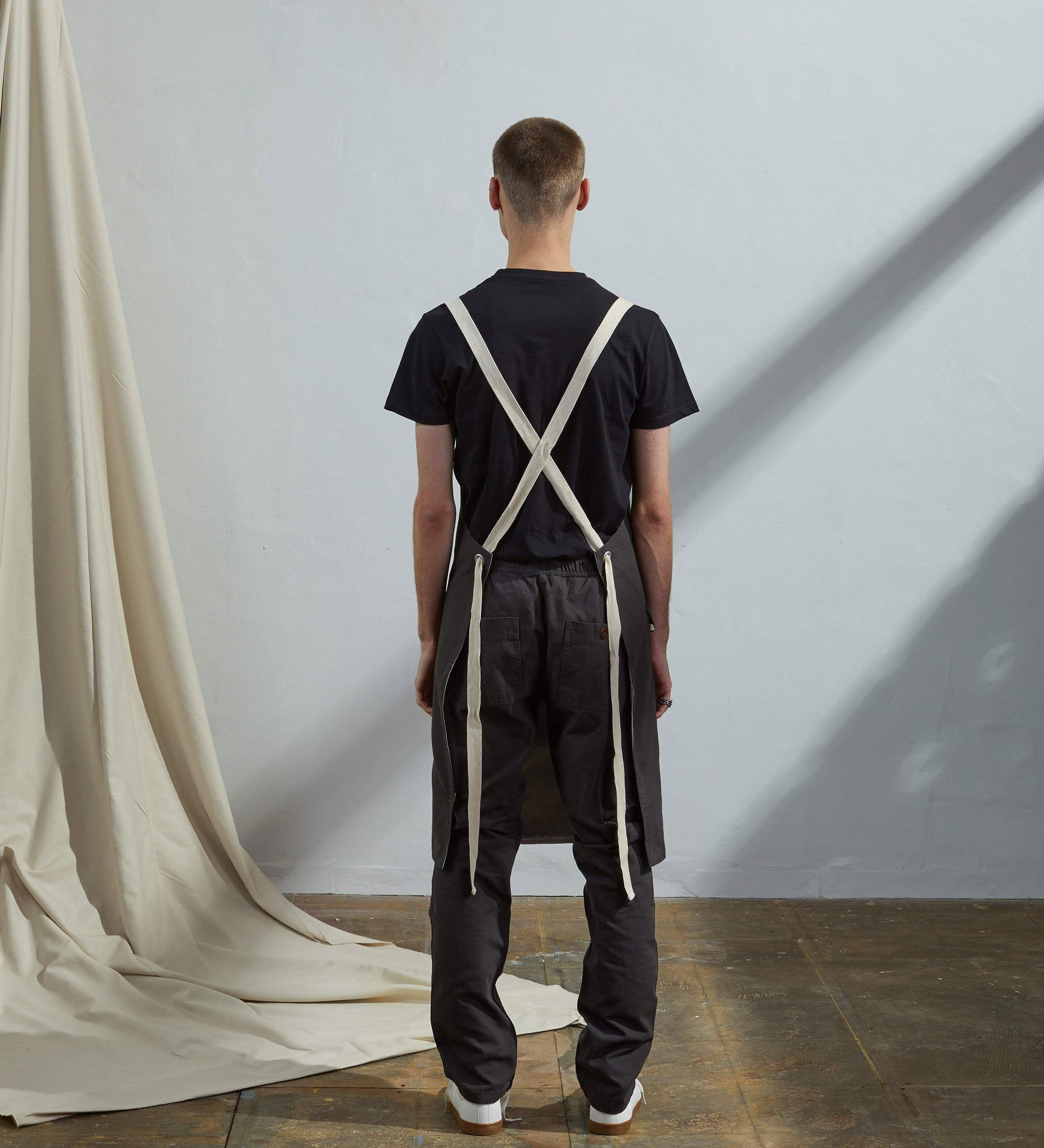 Full-length, rear view of model wearing Uskees #9001 faded black work apron by Uskees. Clear view of cream rear ties.