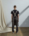 Full-length, rear view of model wearing Uskees #9001 faded black work apron by Uskees. Clear view of cream rear ties.