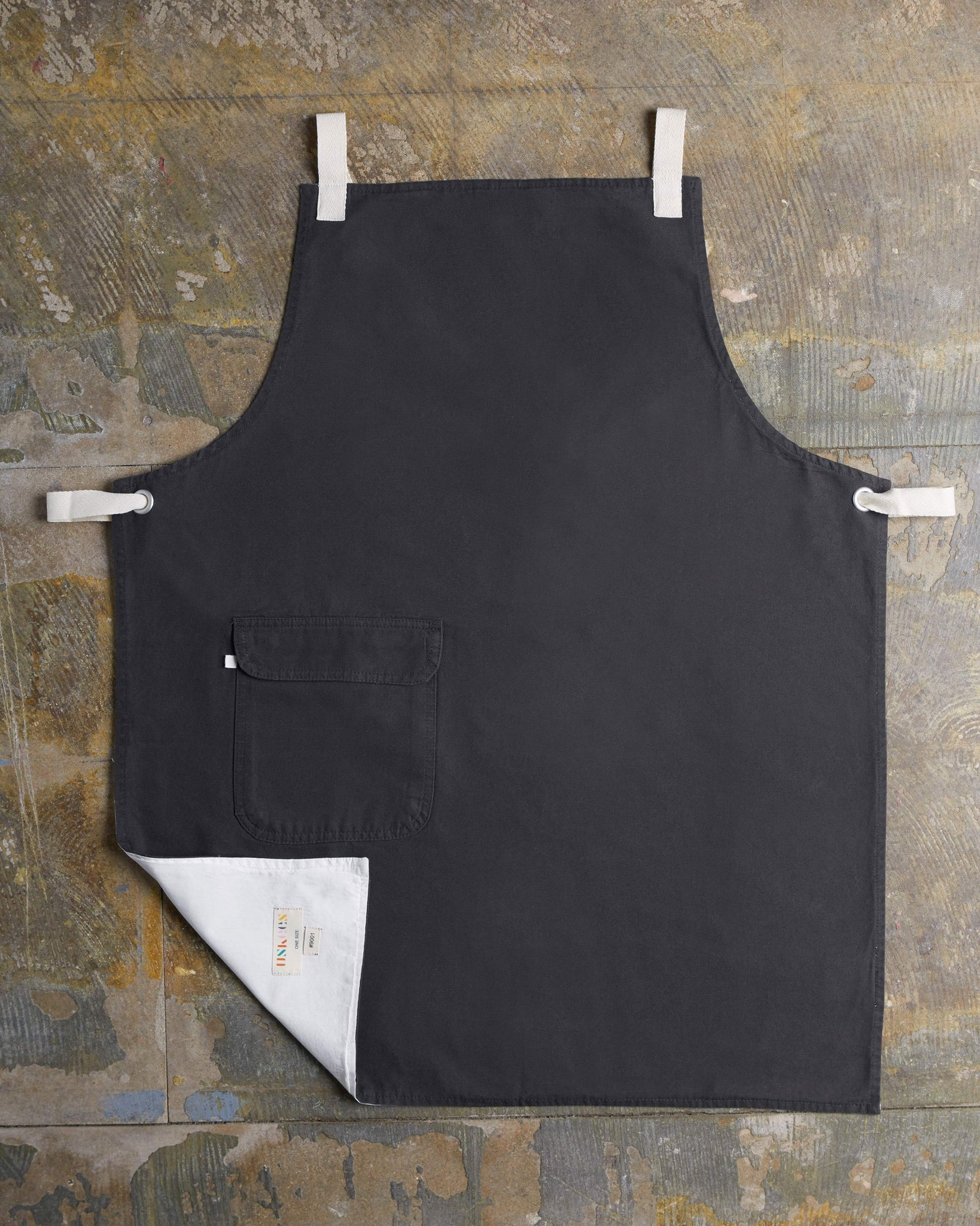 Full, flat view of faded black #9001 work apron by Uskees. Showing hip pocket with left corner folded up to reveal lining and 'Uskees' label.