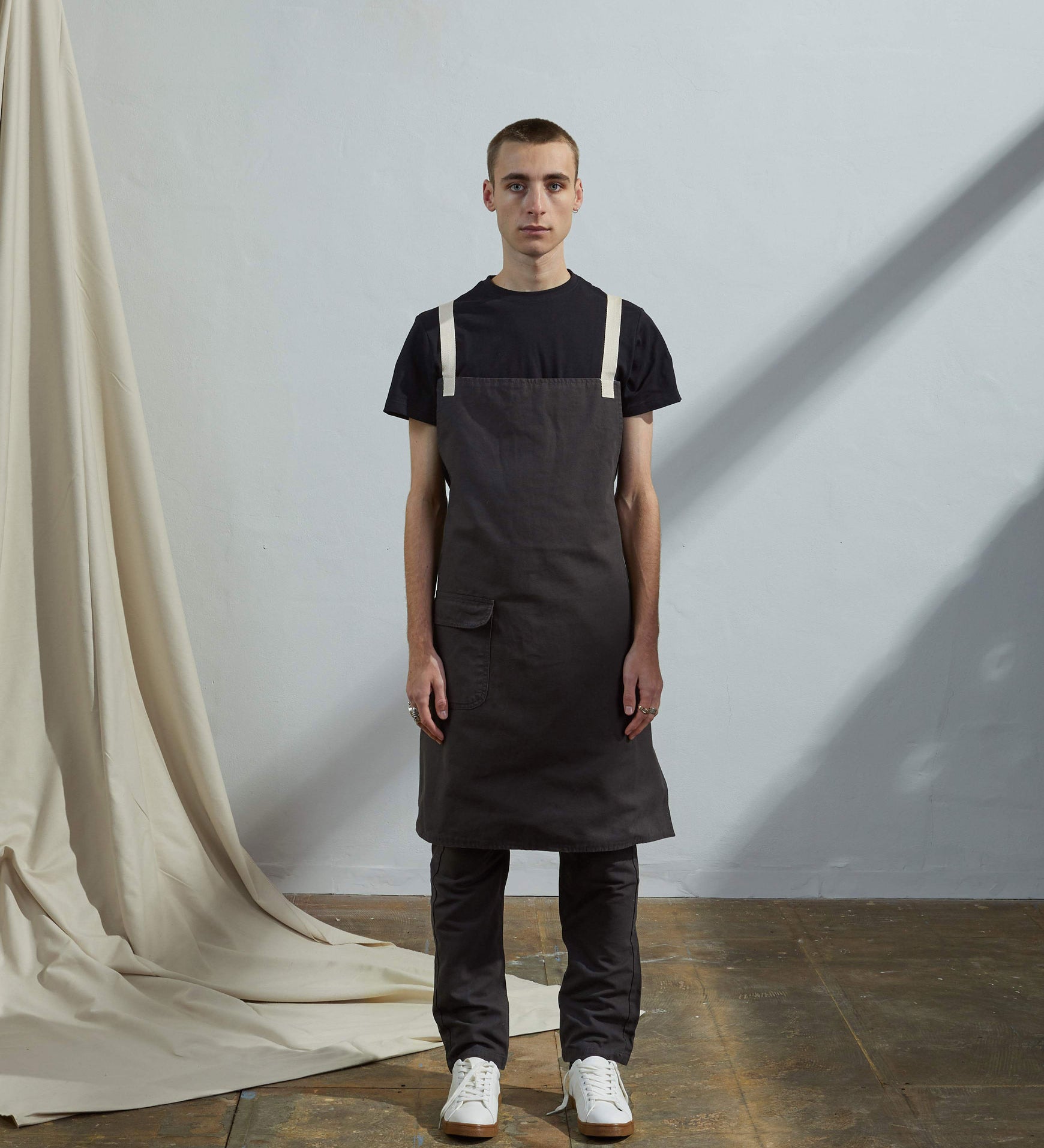 Full-length, front view of male model wearing Uskees #9001 faded black work apron by Uskees. Showing convenient hip pocket and contrasting cream straps.