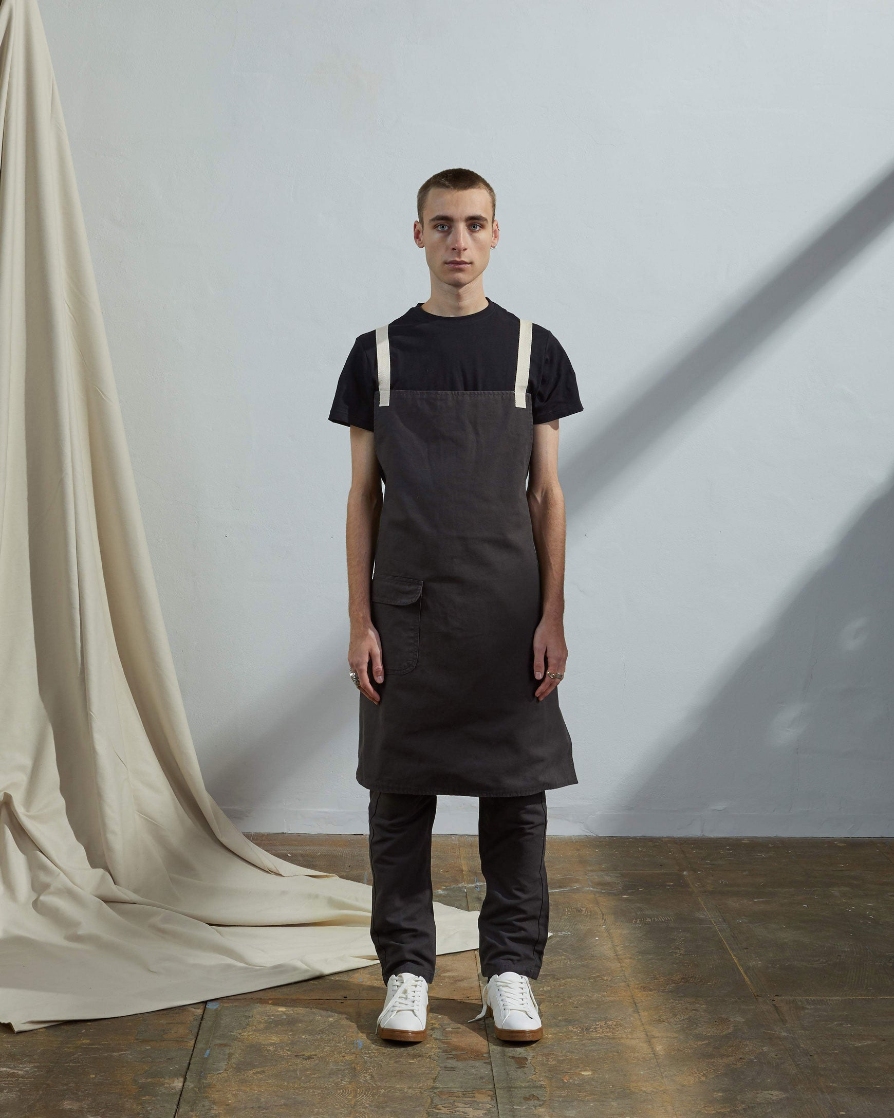 Full-length, front view of male model wearing Uskees #9001 faded black work apron by Uskees. Showing convenient hip pocket and contrasting cream straps.