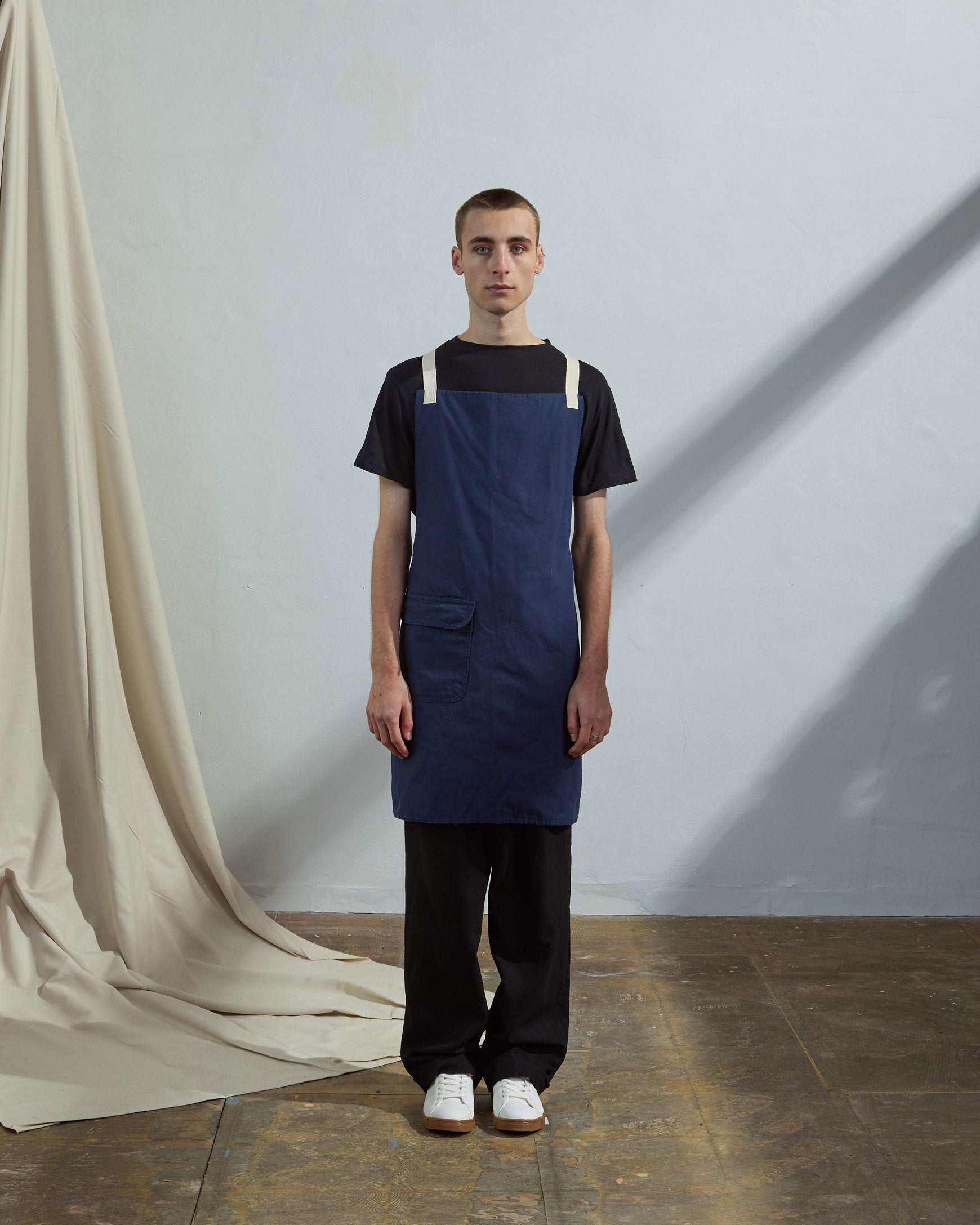 Full-length, front view of male model wearing Uskees #9001 navy work apron by Uskees. Showing convenient hip pocket and contrasting cream straps.