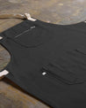 Angled flat view of faded black #9004 carpenter apron by Uskees. Showing pen, phone and pouch pockets and clearer view of organic cotton fabric texture.