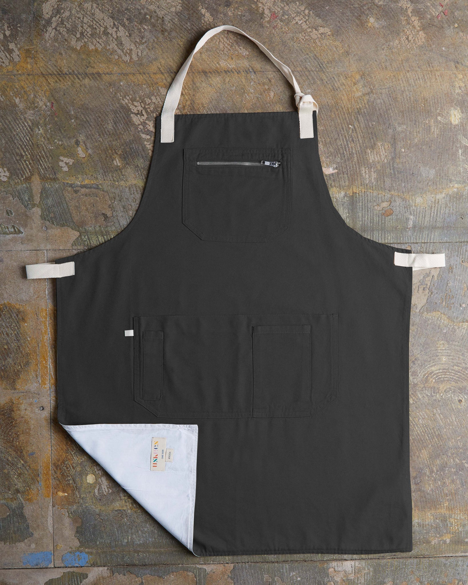 Full, flat view of 'faded black' #9004 carpenter apron by Uskees. Showing pen, phone and pouch pockets with left corner folded up to reveal lining and 'Uskees' label.