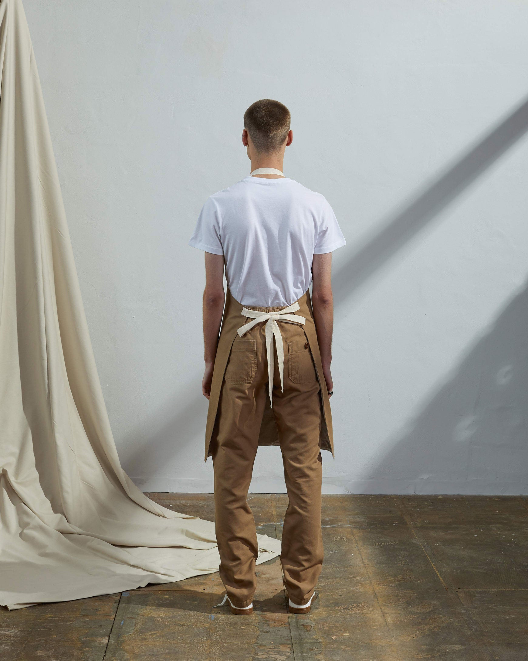 Full-length, rear view of model wearing Uskees #9004 khaki work apron by Uskees. Clear view of cream rear ties.