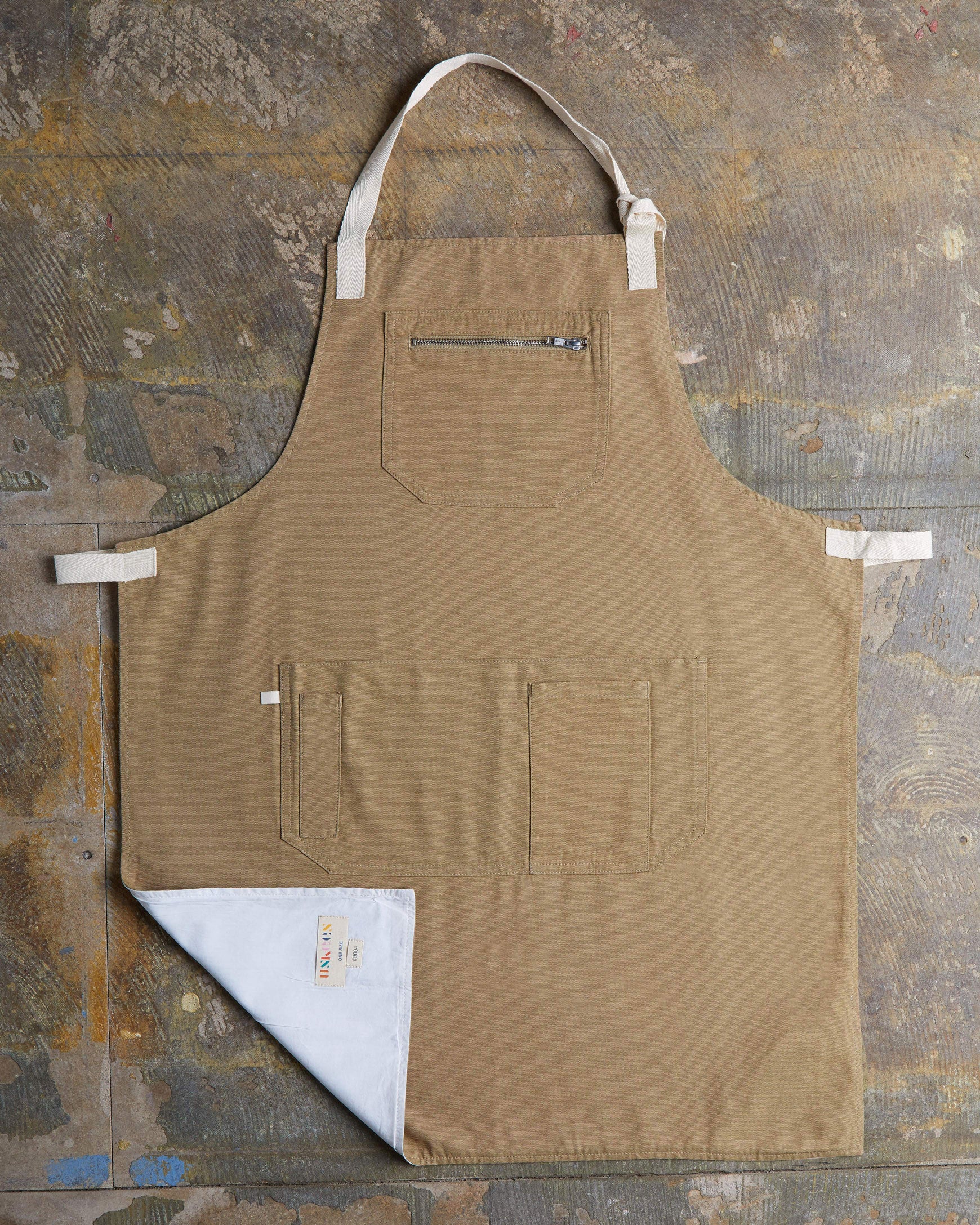 Full, flat view of khaki #9004 carpenter apron by Uskees. Showing pen, phone and pouch pockets with left corner folded up to reveal lining and 'Uskees' label.