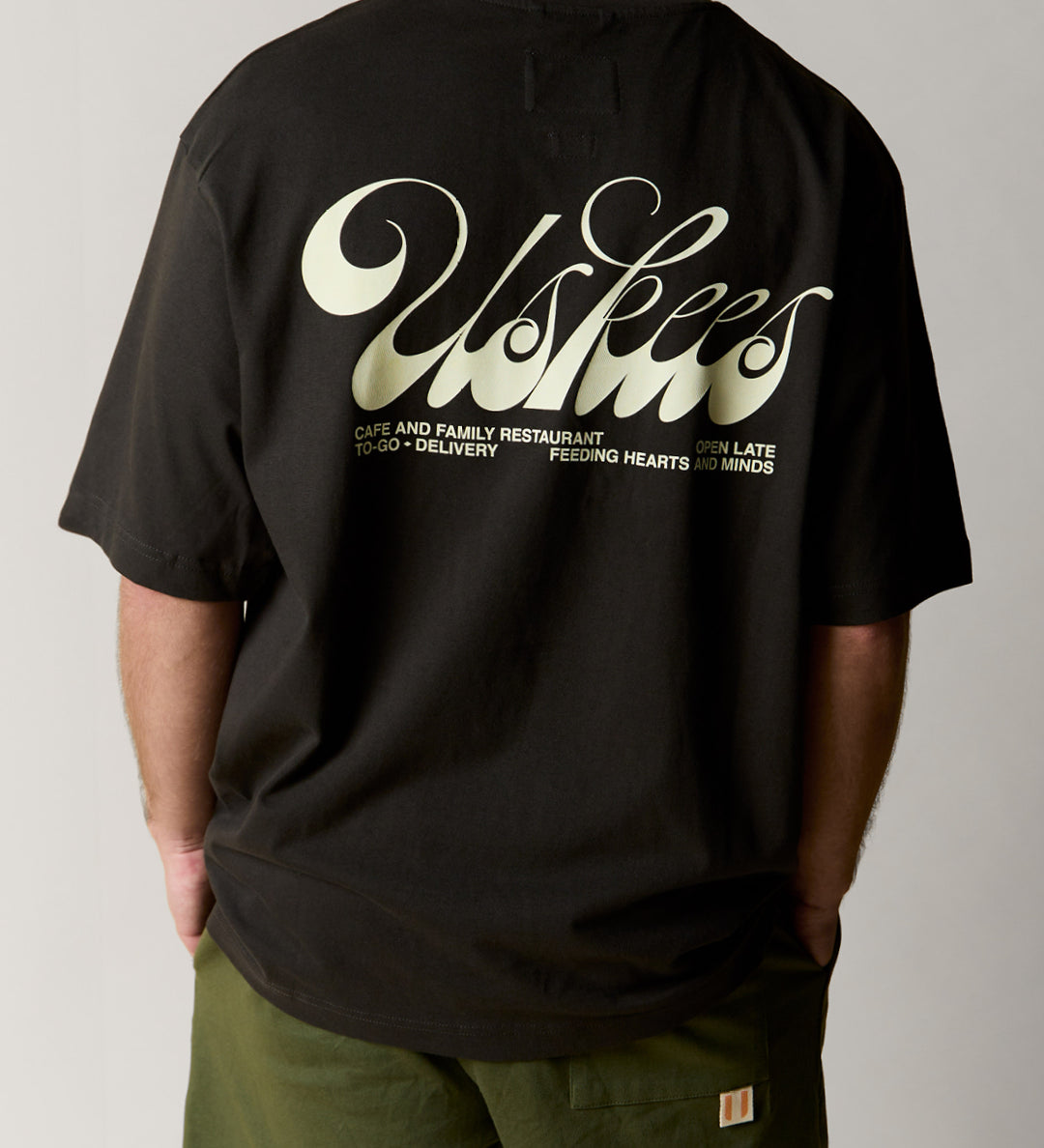back view of model wearing the uskees faded black baggy fit  graphic Tee for men showing the 'Diner' design in cream