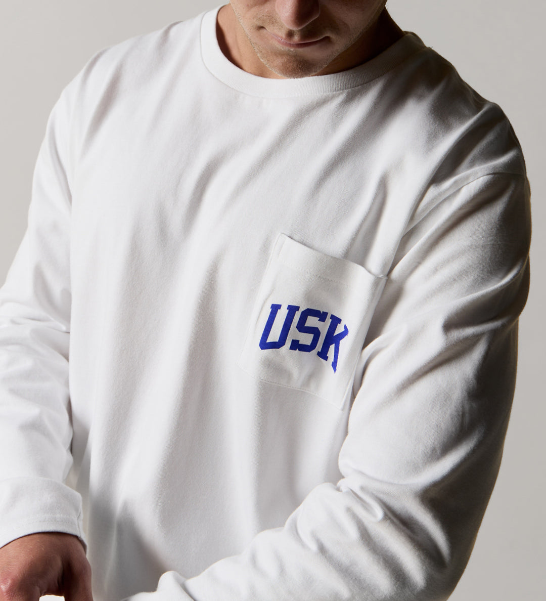 Close shot of model wearing the uskees men's long sleeve graphic  Tee in white showing the blue USK logo on the chest pocket