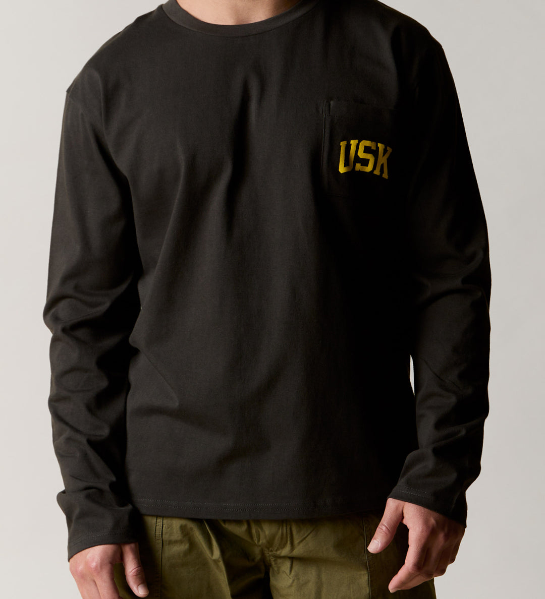 Model wearing the uskees faded black long sleeved Tee for men showing the yellow USK logo on the chest pocket