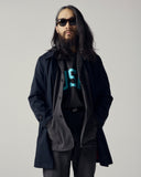 Male model wearing sunglasses and a dark coat over a charcoal cotton drill shirt and Uskees signature t-shirt on a plain background