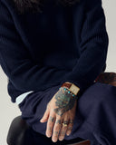 Male model wearing an Uskees dark blue crew neck sweater and pants with tattoos on hands, sitting on a chair.