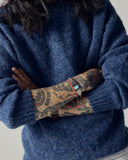 Male model wearing an Uskees 7021 sapphire blue sweater with tattoos on arms and bracelets on a neutral background.